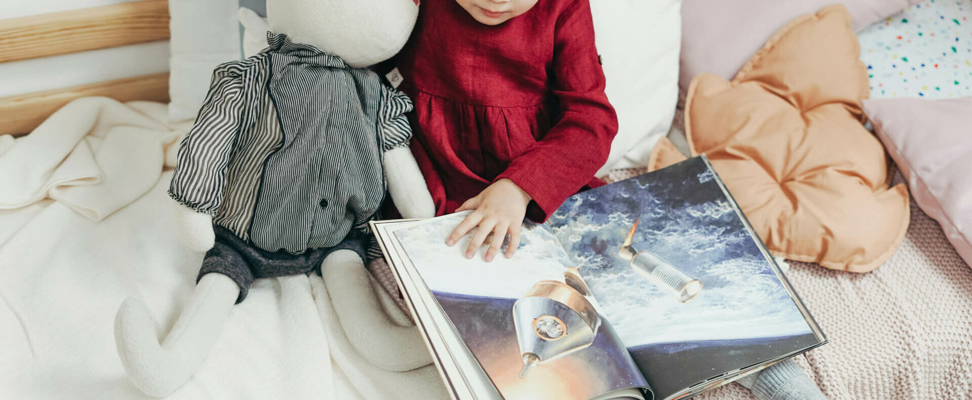 A young girl in a red dress sits on a bed with a stuffed rabbit, looking at a picture book open to a page showing spacecraft in space. Pillows and blankets are around them.