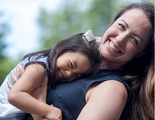 A woman smiles while holding a young girl who is laughing and hugging her. Both appear happy, and the background is softly blurred with greenery.