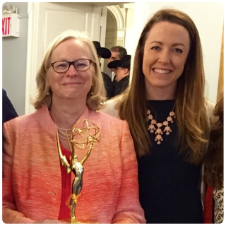 Two women smile at the camera; the woman on the left wears glasses and holds an Emmy award, while the woman on the right, wearing a dark top and a statement necklace, stands beside her. People are visible in the background.
