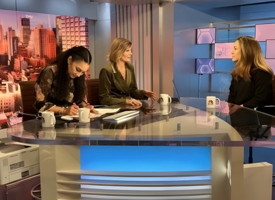 Three women are seated at a curved news desk in a modern TV studio. One woman is writing, another is speaking, and the third listens attentively. Cityscape screens and studio lights are visible in the background.