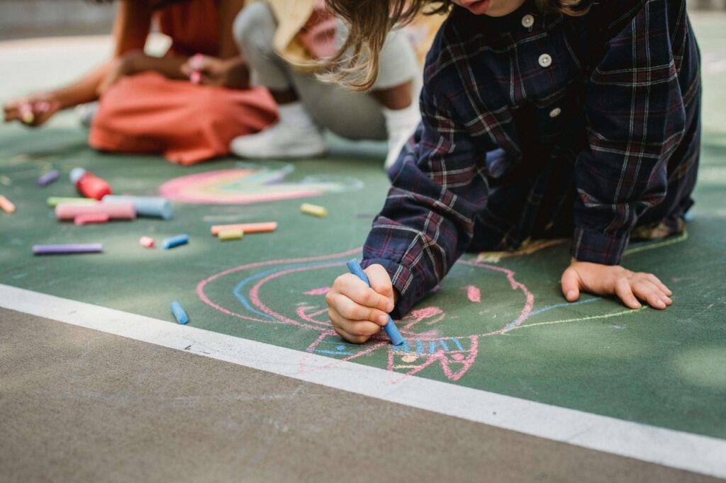 A child draws a colorful cat with chalk on the pavement, surrounded by other chalk drawings and scattered chalk pieces, while another child sits in the background.