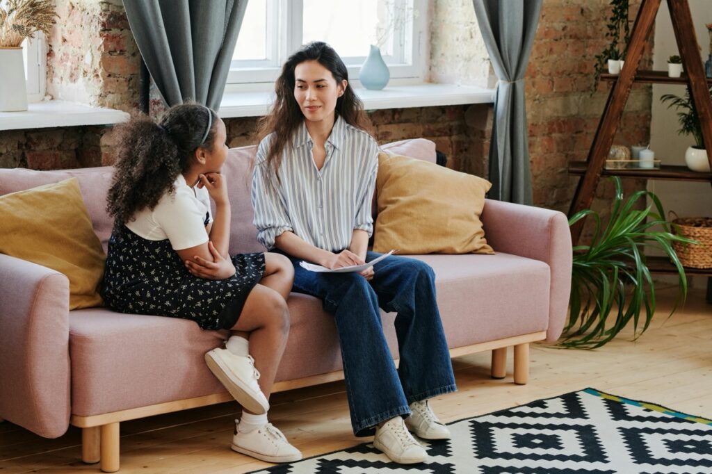 A woman and a young girl sit on a pink sofa in a cozy living room, engaged in conversation. The girl wears a white shirt and floral dress, while the woman holds a notebook and wears a striped shirt and jeans.