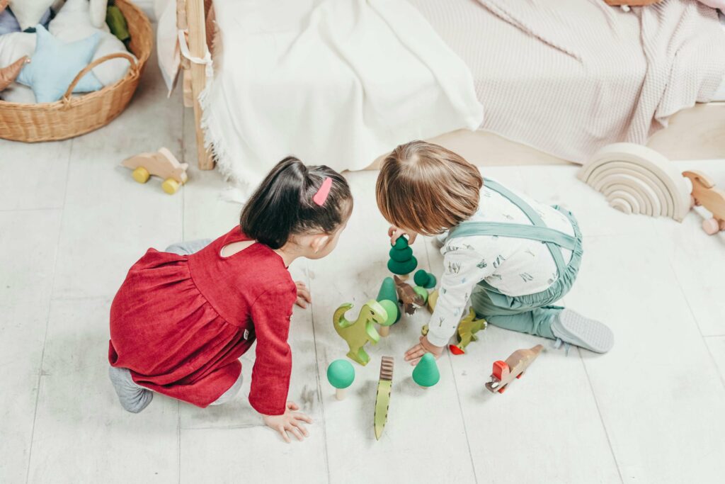 Two young children, one in a red dress and one in overalls, are sitting on the floor playing with wooden toy trees and animals next to a bed in a cozy, light-filled room.
