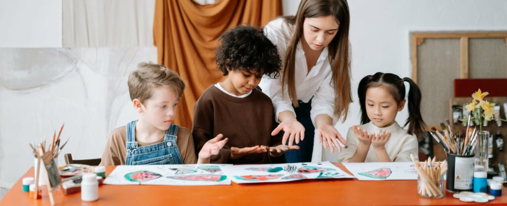 A teacher helps three young children examine paint on their hands as they sit at a table covered with colorful artwork and painting supplies.