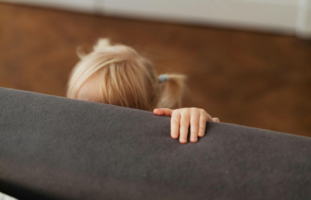 A young child with blonde hair peeks over the back of a gray sofa, with only the top of their head and one hand visible. The background is blurred.