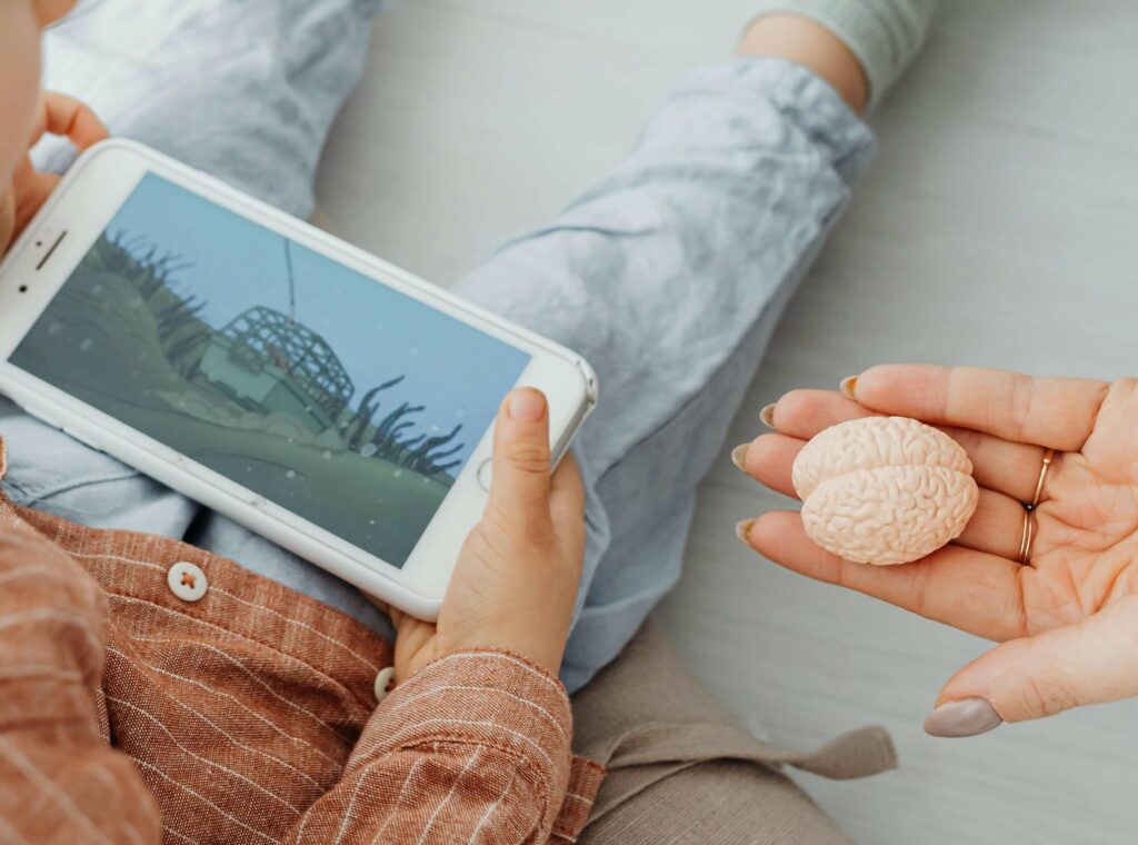 A child holding a smartphone with an animated video on screen, while an adult hand shows a small model of a brain. The child is sitting on the floor, wearing light blue pants and a brown shirt.