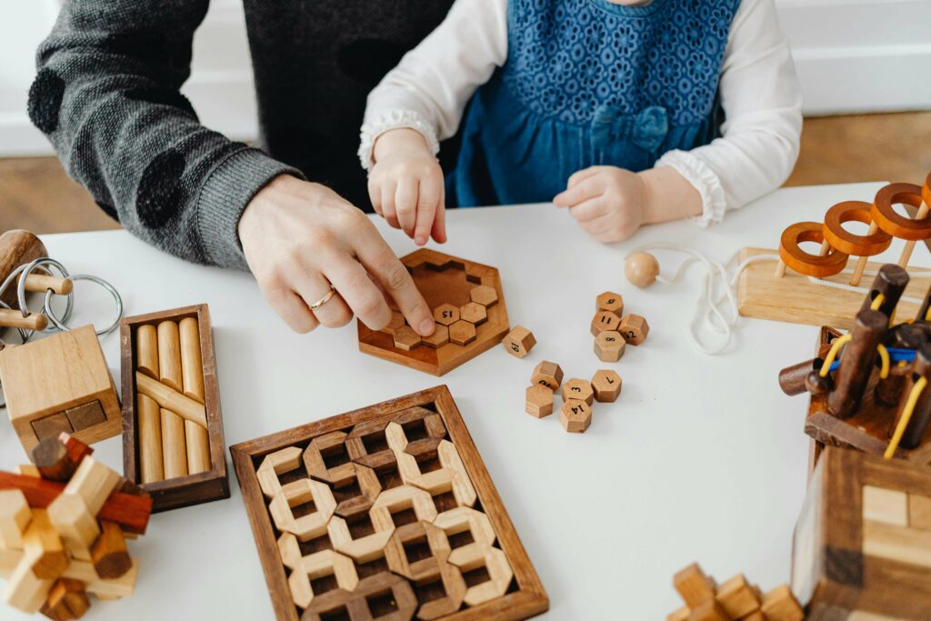 An adult and a child sit at a table playing with various wooden puzzles and geometric toys. The adult’s hand helps arrange hexagonal pieces while the child, dressed in blue, reaches for a piece.