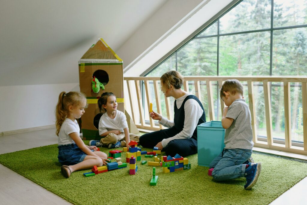 Four children sit on a green rug, playing with colorful building blocks in a bright room with large windows and a sloped ceiling. A playhouse made of cardboard is in the background.