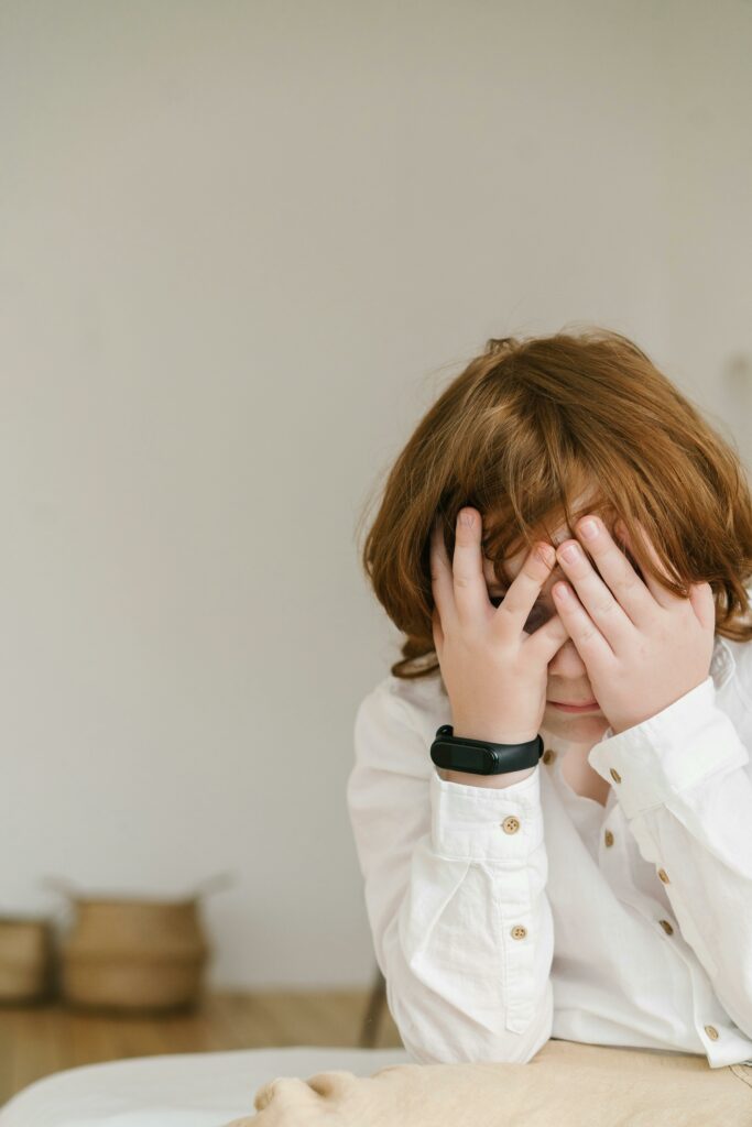 A child with reddish-brown hair, wearing a white shirt and a smartwatch, sits on a bed with their face buried in their hands, appearing upset. The background is minimal, with baskets on the floor.