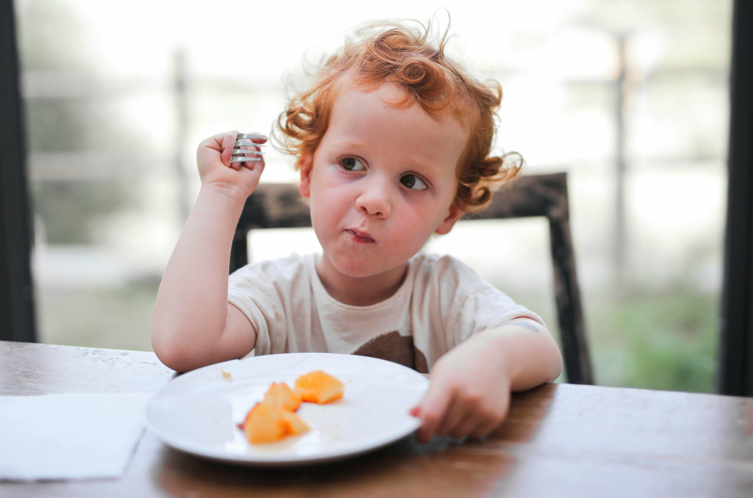 A young child with curly red hair sits at a table, holding a fork and looking to the side while eating pieces of fruit from a white plate. The background is softly blurred.