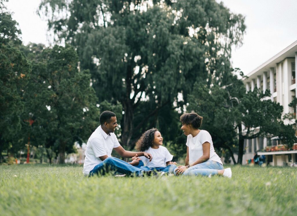 A family of three sits on the grass in a park, smiling and talking together. Trees and a building are visible in the background, and all three wear white shirts and jeans.