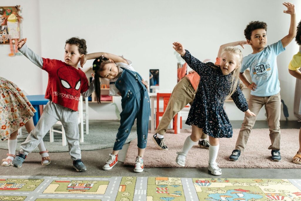 A group of young children stand in a classroom, smiling and stretching to the side with one arm raised, participating in a playful group activity on a colorful carpet.