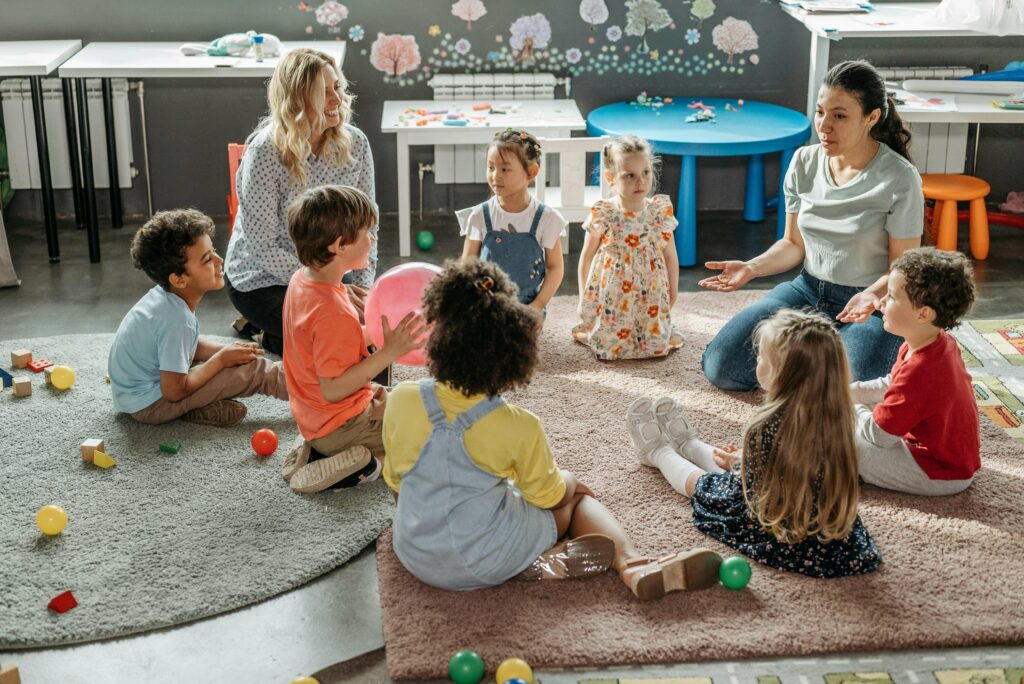 A group of young children sit in a circle on a rug in a classroom, listening to two adults. Toys and colorful balls are scattered around, and there are tables and drawings on the walls in the background.