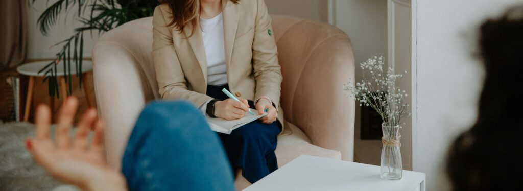 A person in a beige blazer sits in a chair, writing in a notebook, while another person faces them with an outstretched hand. A small table with a vase holds white flowers in the foreground.