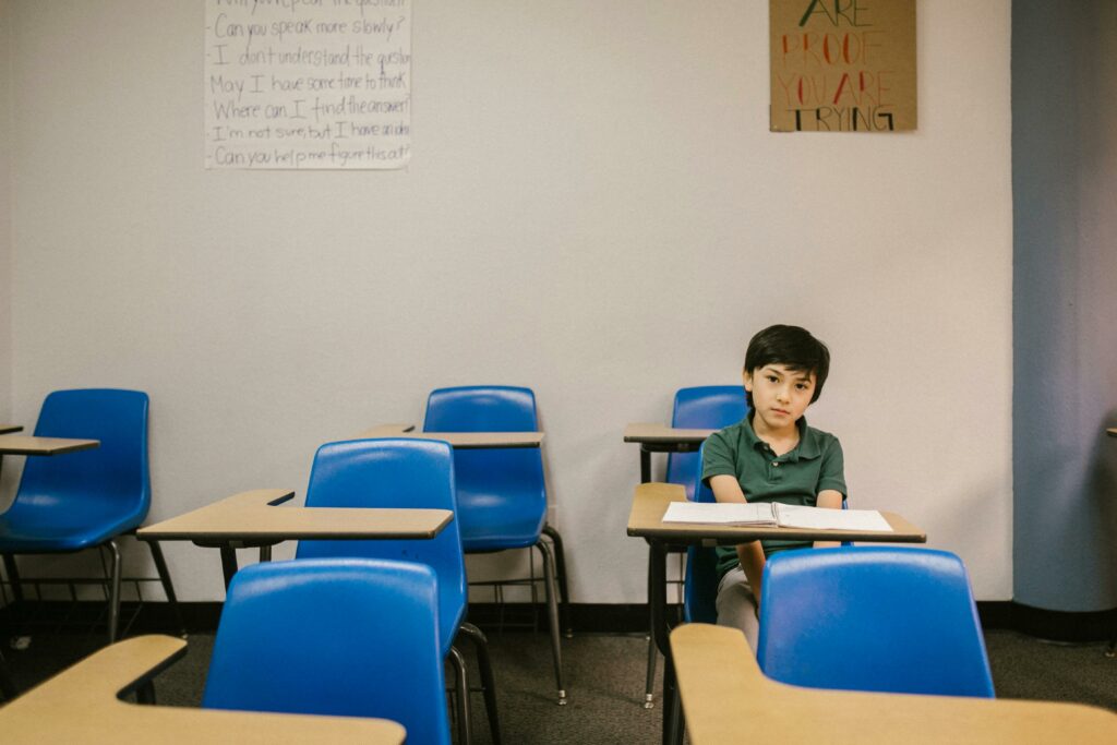 A young boy sits alone at a desk in a mostly empty classroom with blue chairs. Handwritten notes are on the wall, and a motivational sign hangs above. The boy looks thoughtful and is facing the camera.
