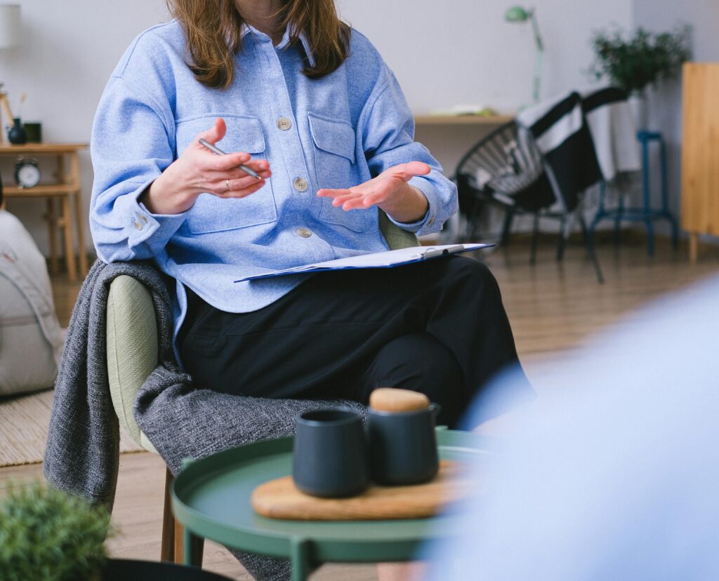 A person in a blue shirt sits in a chair gesturing with their hands, holding a clipboard on their lap. In the foreground, a green table holds two black cups and a small wooden tray. The setting appears to be a cozy room.