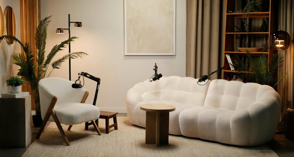 A cozy podcast studio with a white armchair, a plush white sofa, a small round wooden table, microphones on stands, potted plants, a bookshelf, and warm ambient lighting.