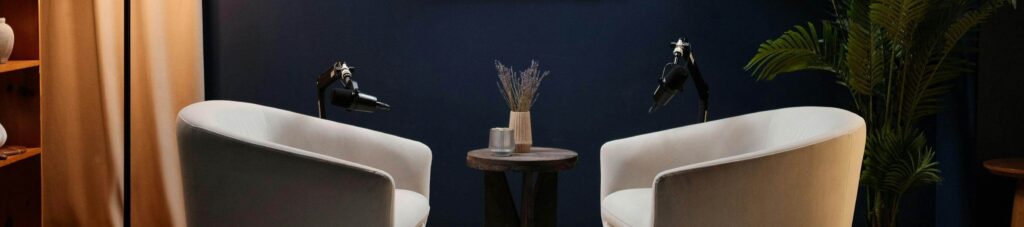 Two white armchairs face each other with microphones positioned in front of each, separated by a small round table holding a vase with dried lavender, set against a dark blue wall with a plant in the corner.