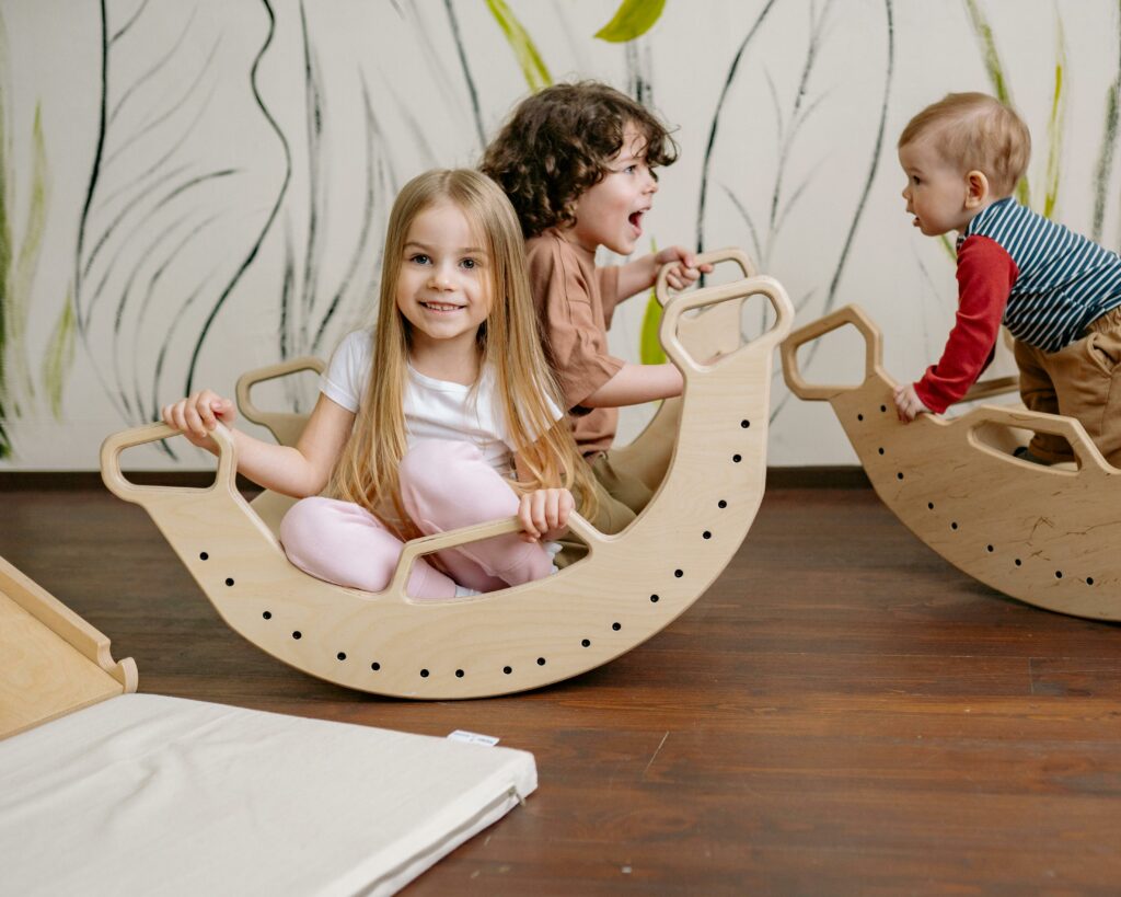 Three young children play indoors on wooden rocking toys. One girl with long hair smiles at the camera, while two other children, a boy and a toddler, are engaged in play. A mural with leaves is on the wall behind them.