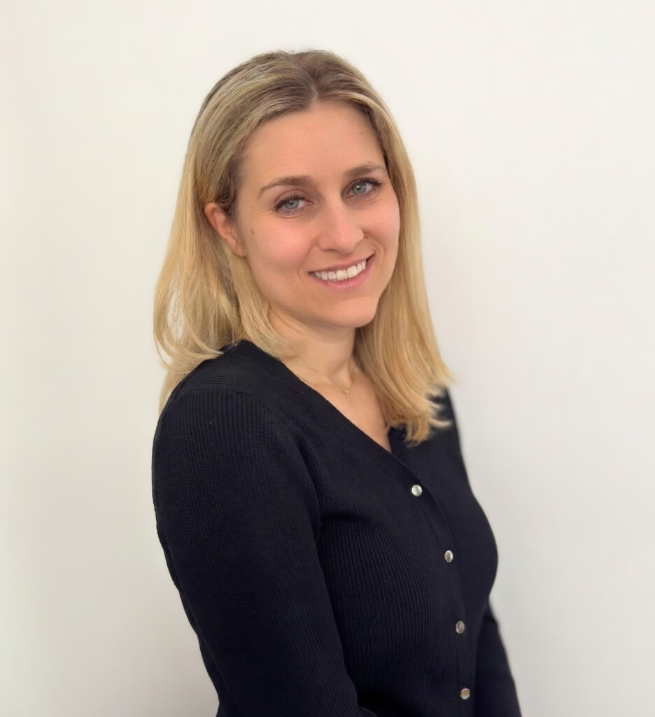 A woman with straight blonde hair and light eyes, wearing a black buttoned sweater, stands against a plain white background and smiles gently at the camera.