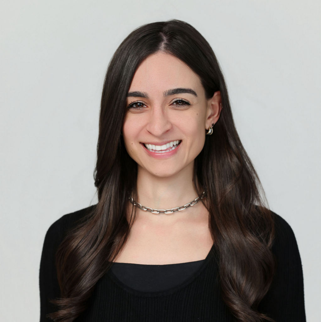 A woman with long brown hair, wearing a black top and silver necklace, smiles at the camera against a plain light background.