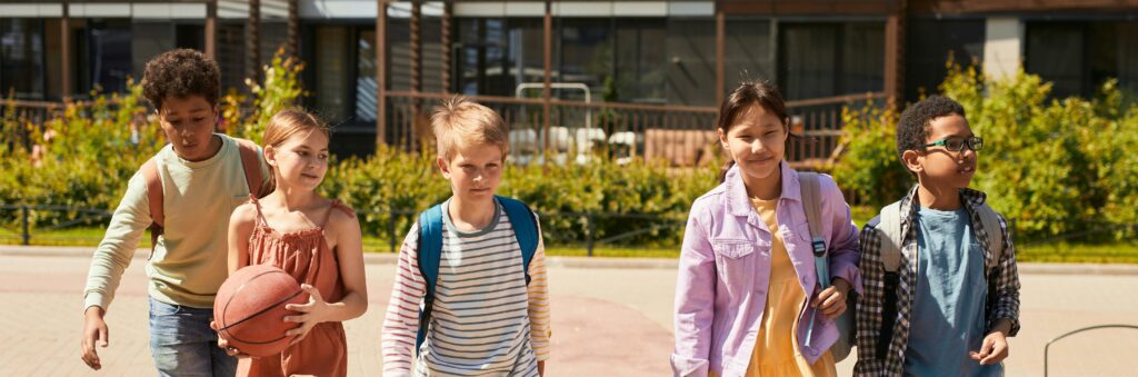 Five children walk together outside, carrying backpacks. One child holds a basketball. They appear to be friends, smiling and talking, with buildings and greenery in the background.