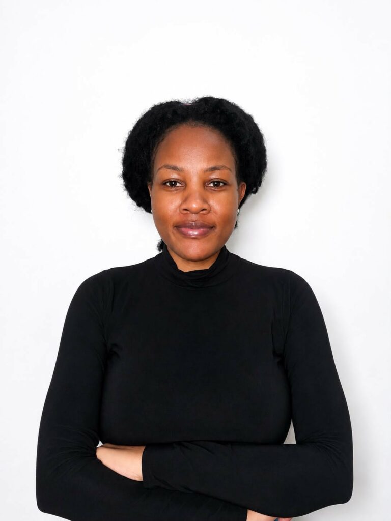 A woman with dark hair styled in two braids, wearing a black long-sleeve top, stands against a plain white background with her arms crossed, looking confidently at the camera.